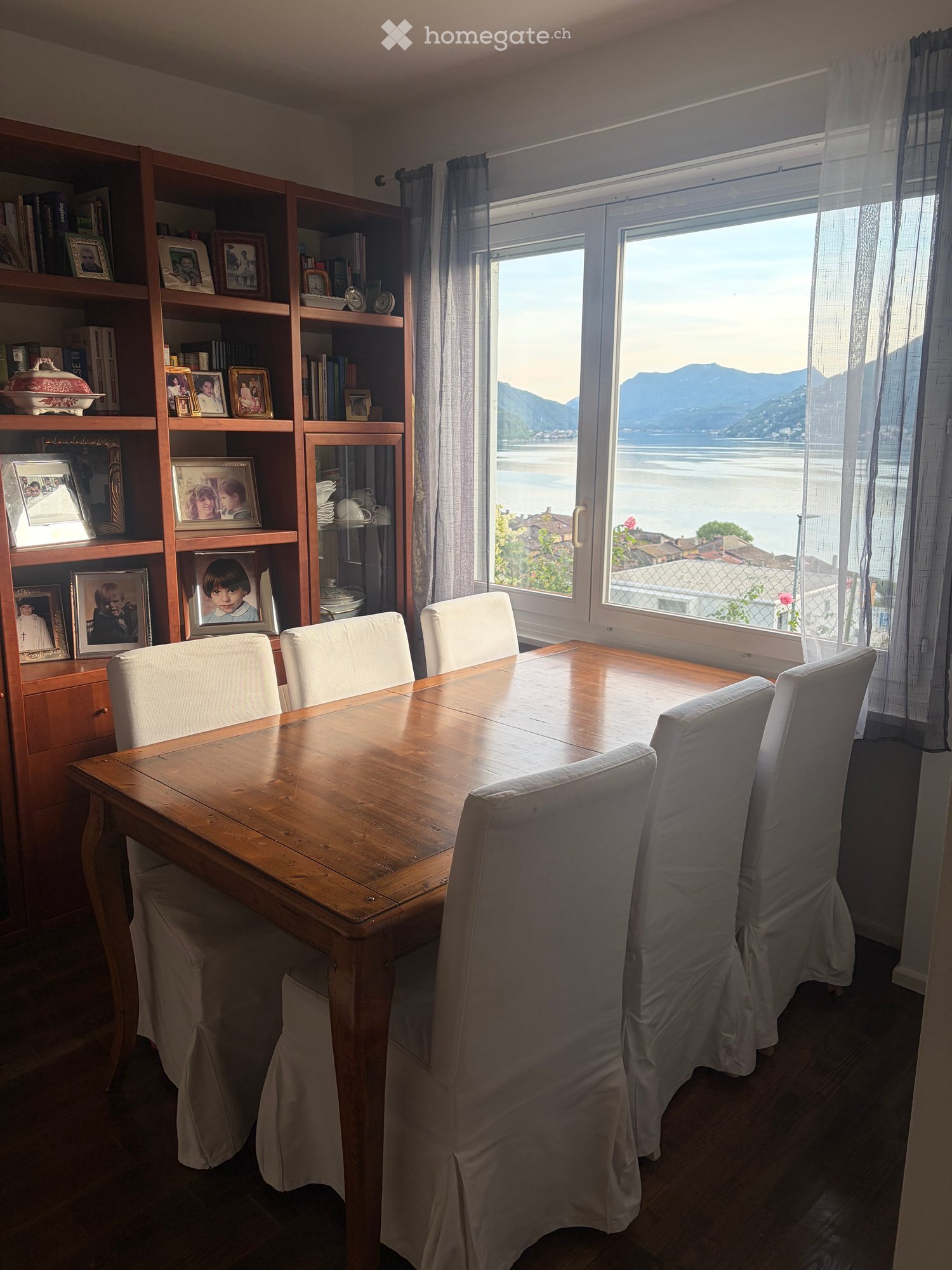 Dining room with a wooden table, six chairs, a large bookshelf filled with books and picture frames, and a scenic view of a lake and mountains through large windows.