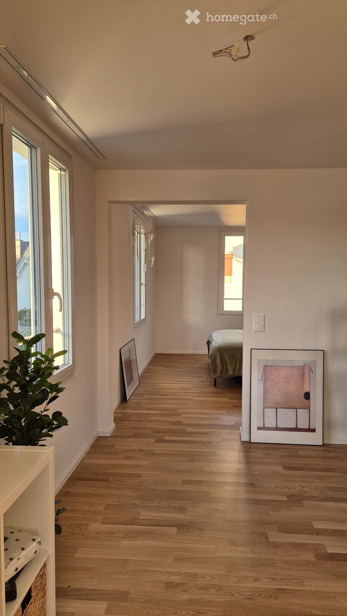 A bright hallway with wooden flooring, large windows, and a view into a bedroom area with a bed and artwork.
