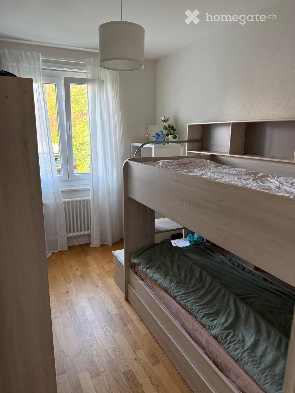 Bright bedroom with wooden floor, large window with white curtains, bunk bed with green bedding, nightstand, and shelf.