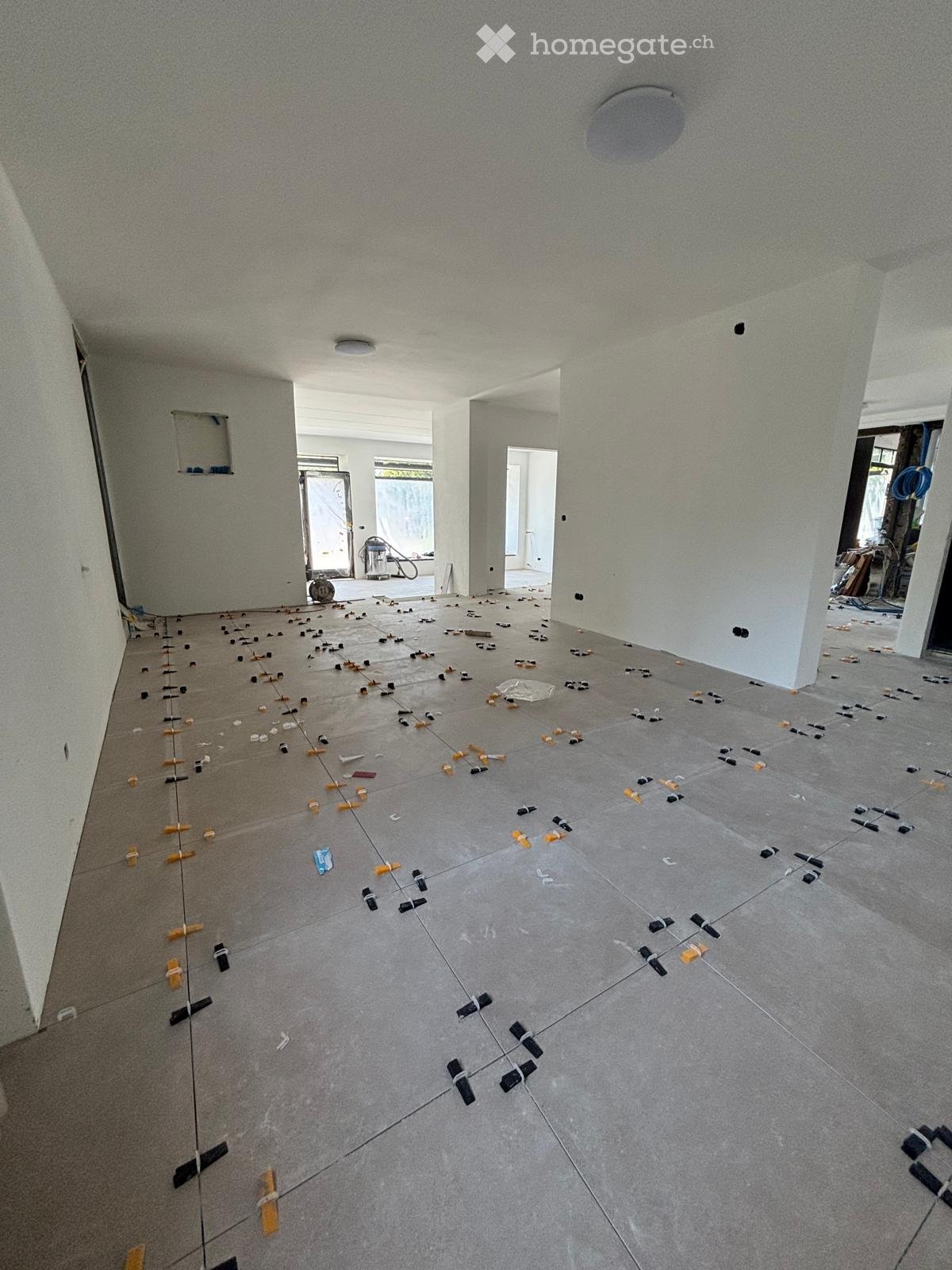 Empty room with grey tiled floor and white walls, showing tile spacers and levelers for installation.