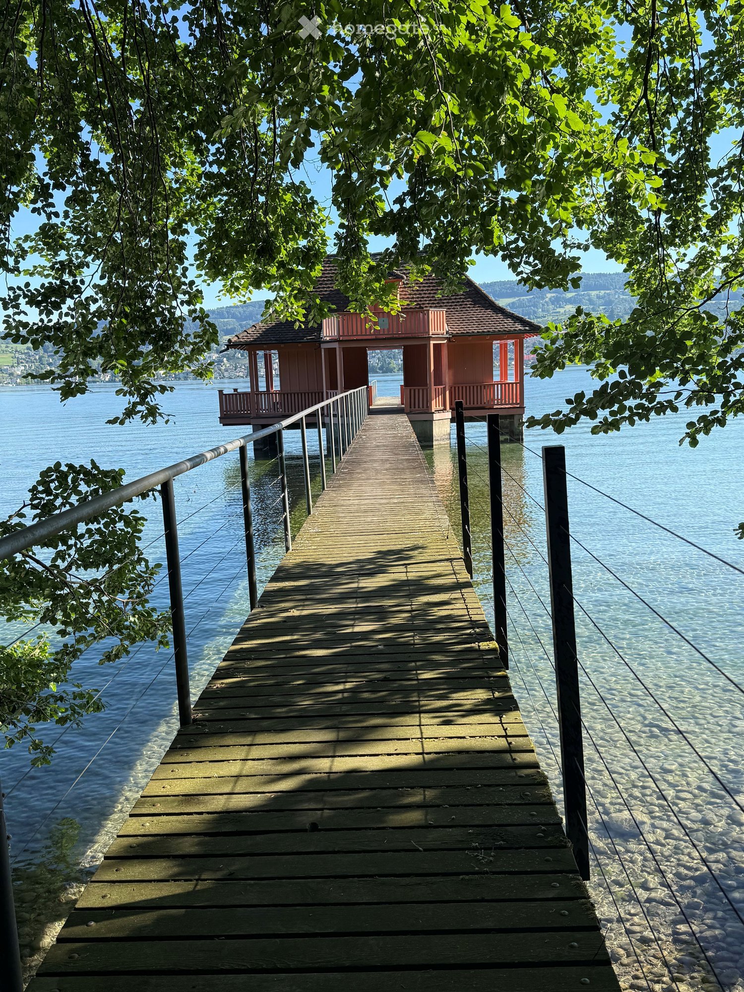 A wooden dock extends into a calm lake, surrounded by lush green trees and a small pink gazebo on the far end.