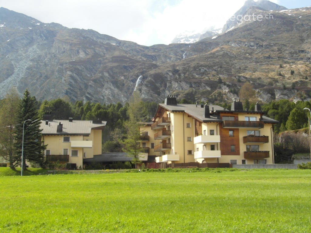 Multi-story residential building with balconies, surrounded by green grass and trees, with a mountain view in the background.
