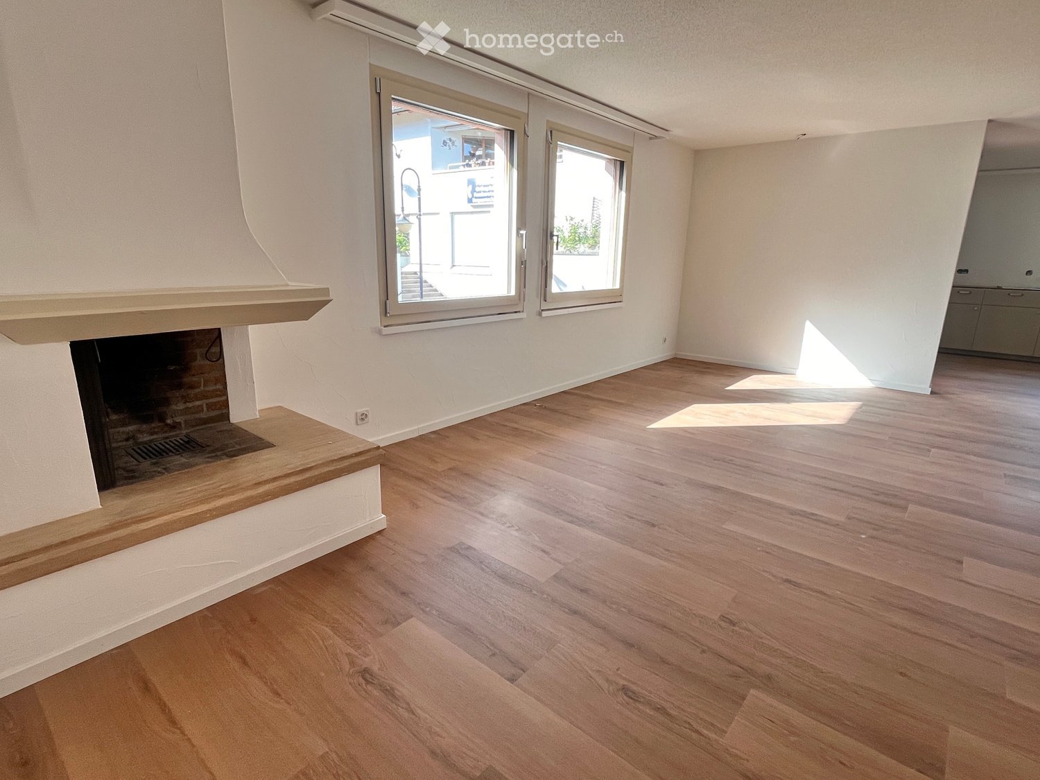 Empty room with light wooden flooring, white walls, and a fireplace with a wooden mantel. Large windows allow natural light to enter.