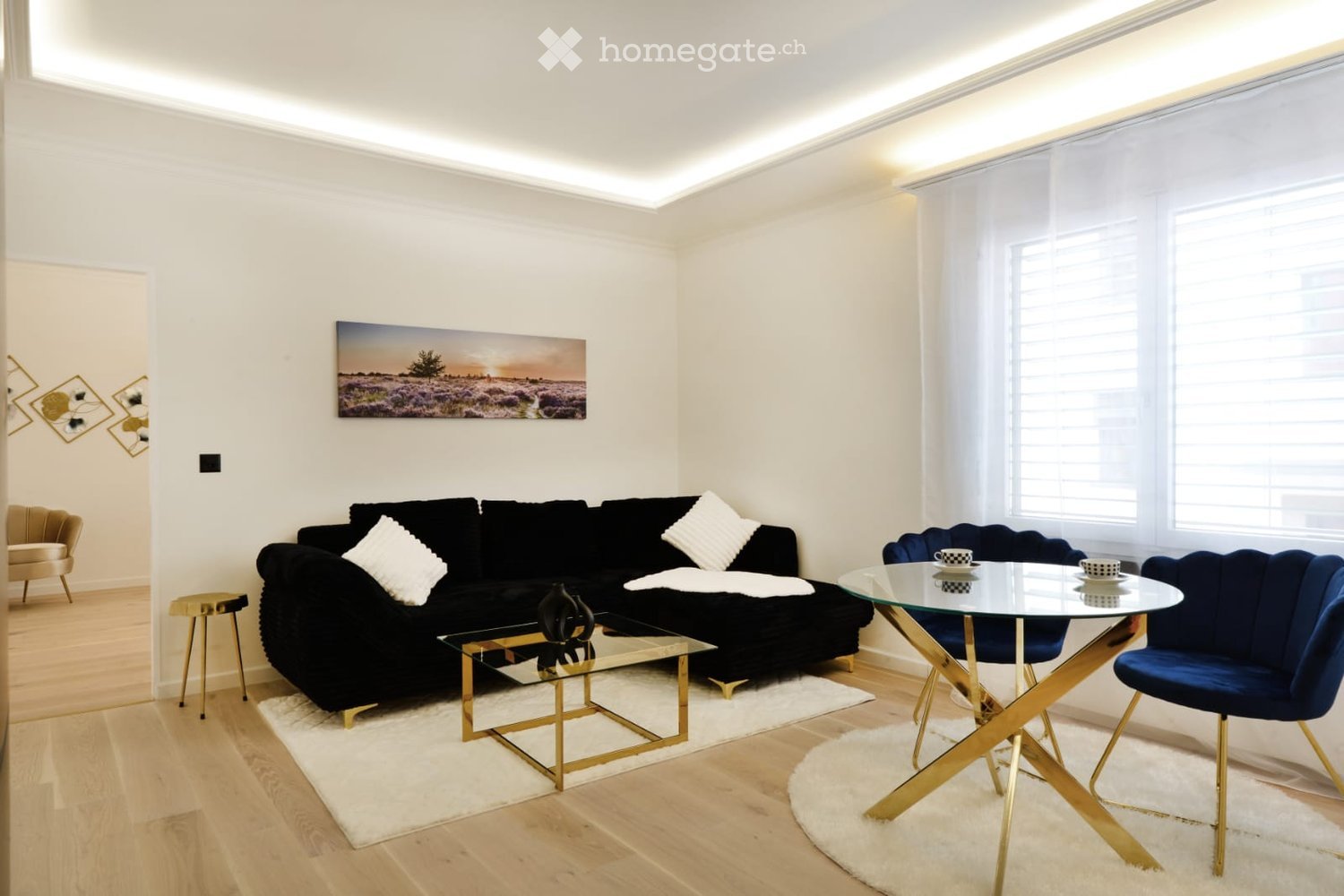 Modern living room with a black sectional sofa, glass coffee table, blue accent chairs, and a glass dining table with matching chairs. The room features light wooden flooring, white walls, and large windows with white curtains.