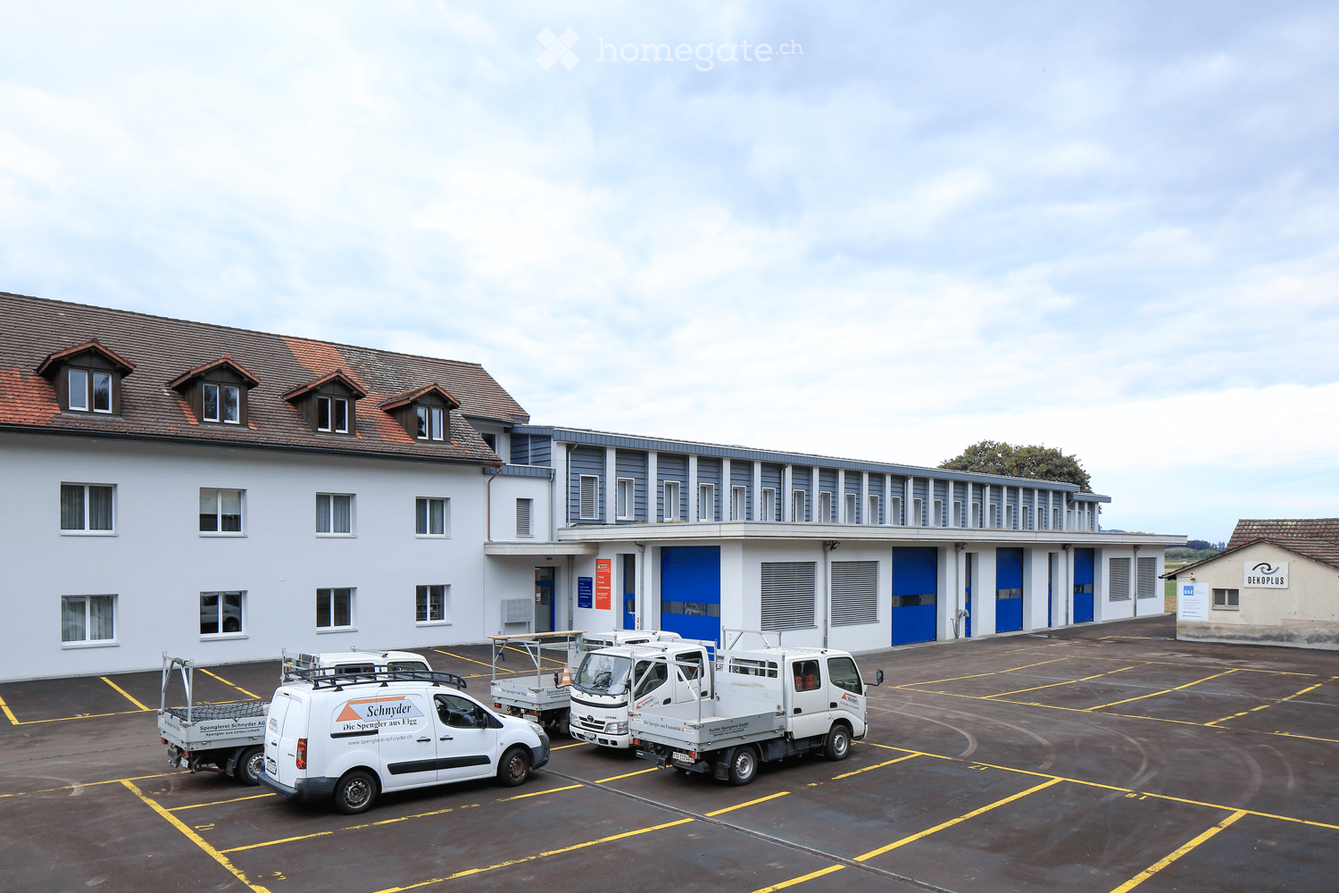 A large industrial-style building with a gray facade and blue garage doors, surrounded by a paved parking lot with several white service vans parked.