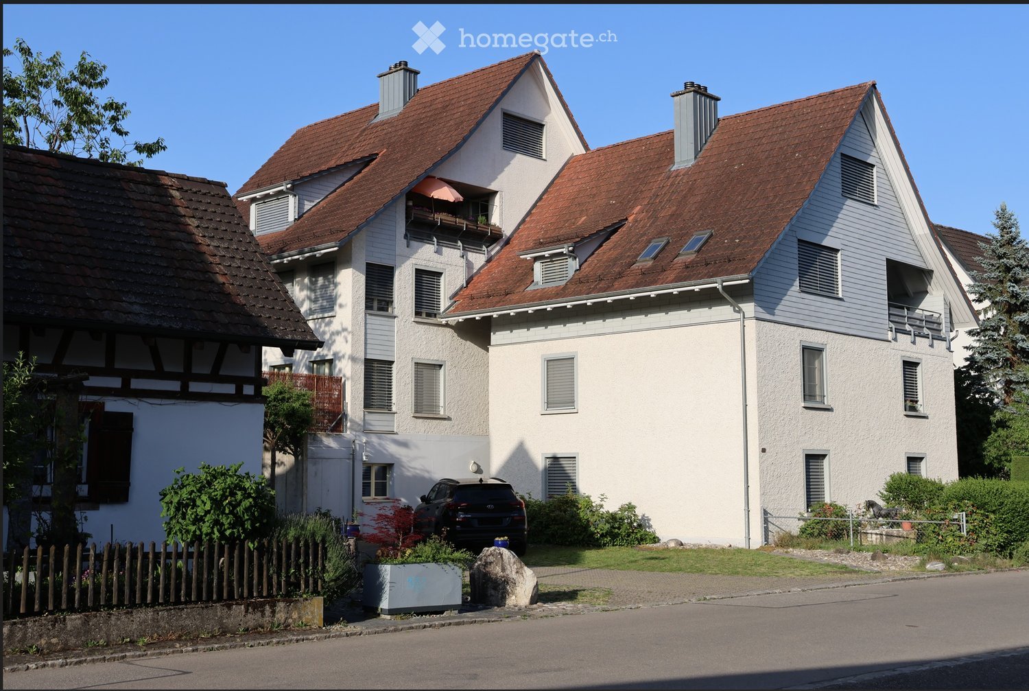 Two-story house with red-tiled roof, white walls, multiple windows, and balconies. A car is parked in the driveway, and there is a small garden area with plants and a wooden fence.