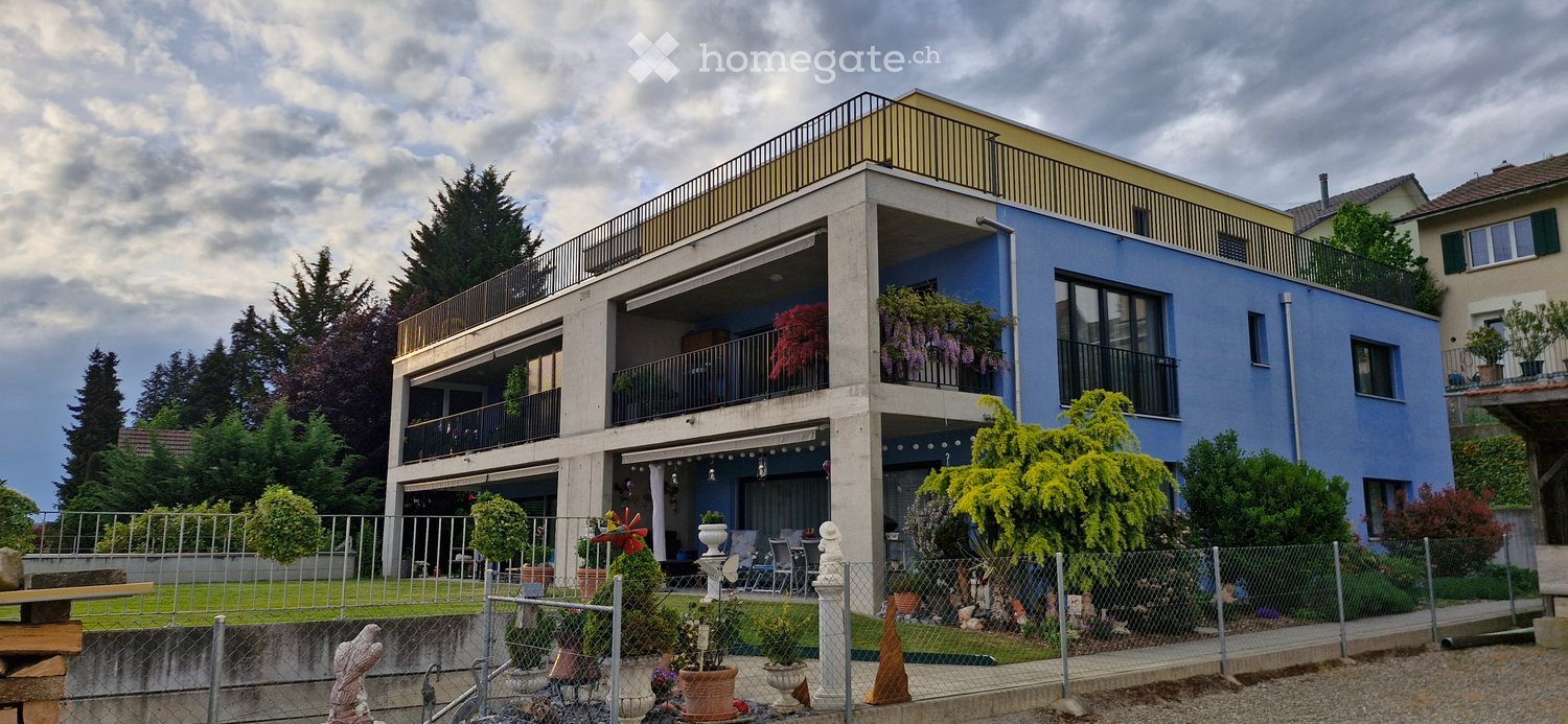 A modern two-story house with a blue exterior and yellow accents. The house has multiple balconies with railings, and a terrace on the top floor. The exterior is surrounded by a garden with various plants and trees.