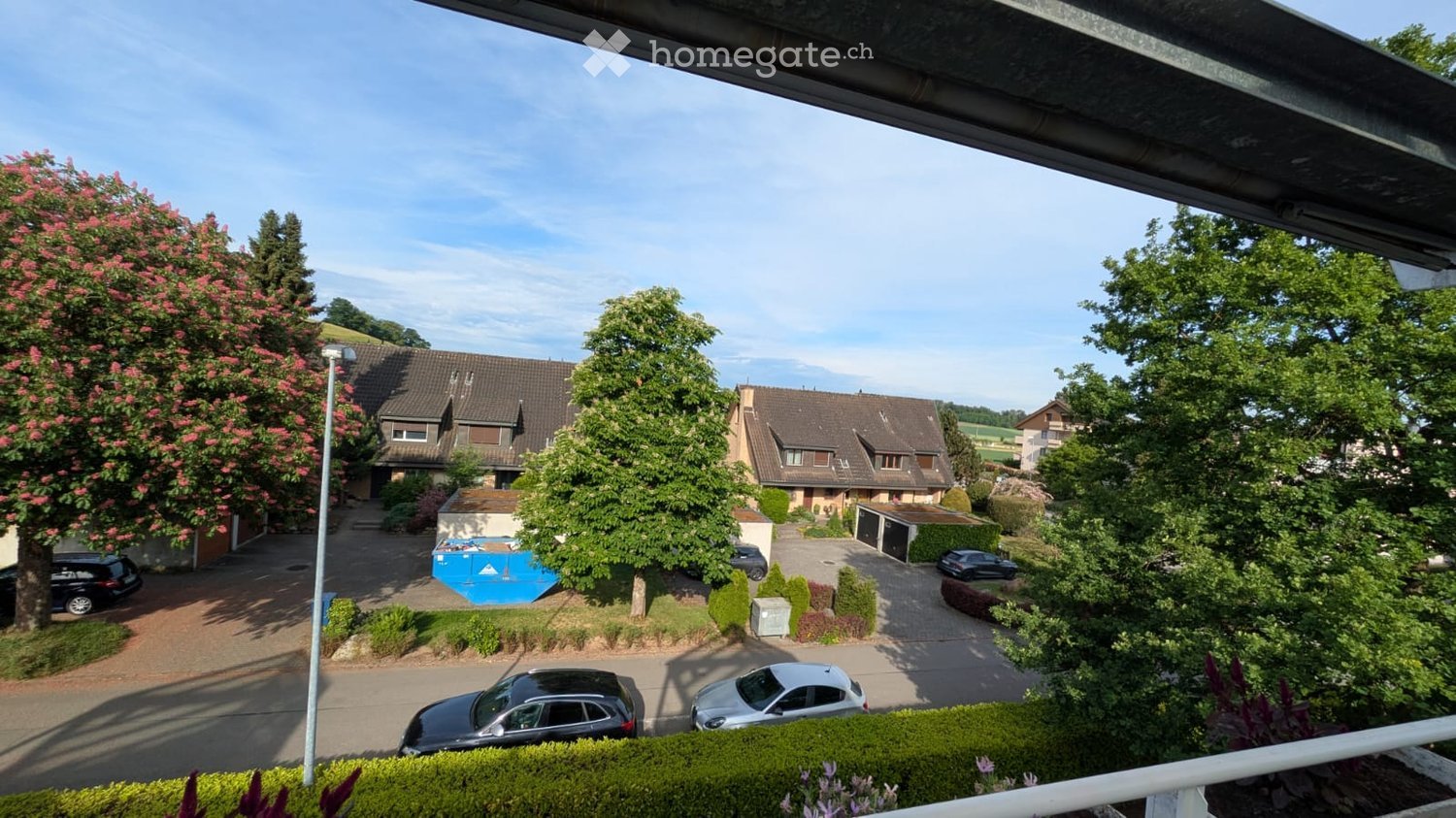 Balcony view of a residential area with houses, parked cars, and greenery.