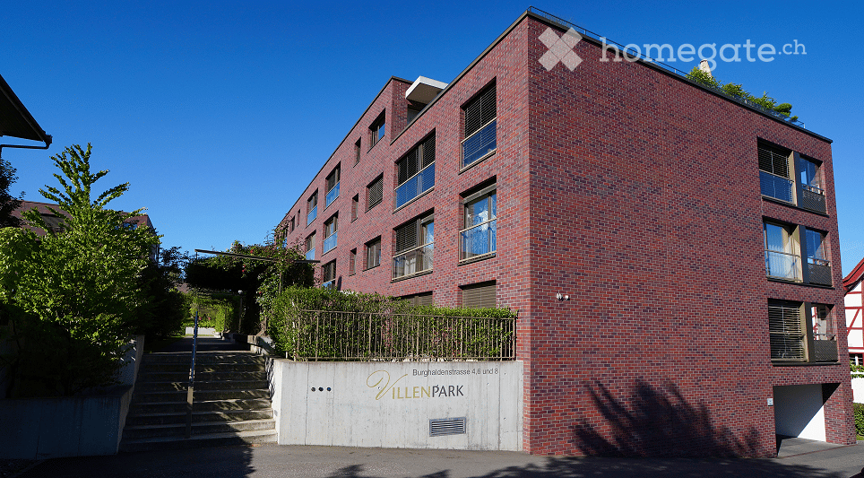 Modern red brick apartment building with multiple floors, balconies, and a raised ground floor entrance.