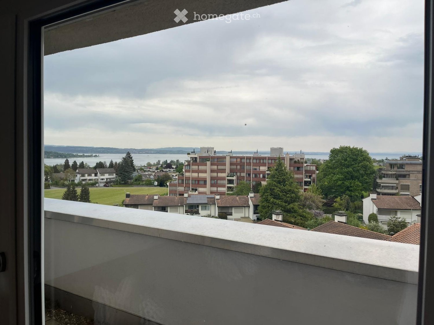 Balcony overlooking a cityscape with buildings, trees, and a body of water in the distance under a cloudy sky.