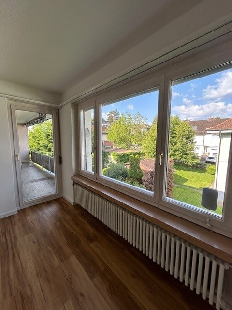 Bright, empty room with wooden flooring, large windows offering a view of a green garden, and a balcony accessible through a glass door.
