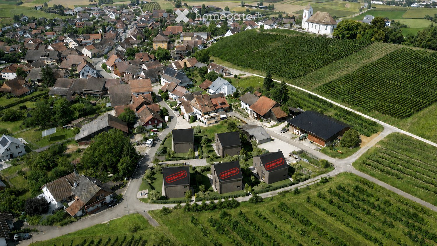 Aerial view of a small village with houses, vineyards, and a church, surrounded by green fields and rural landscape.