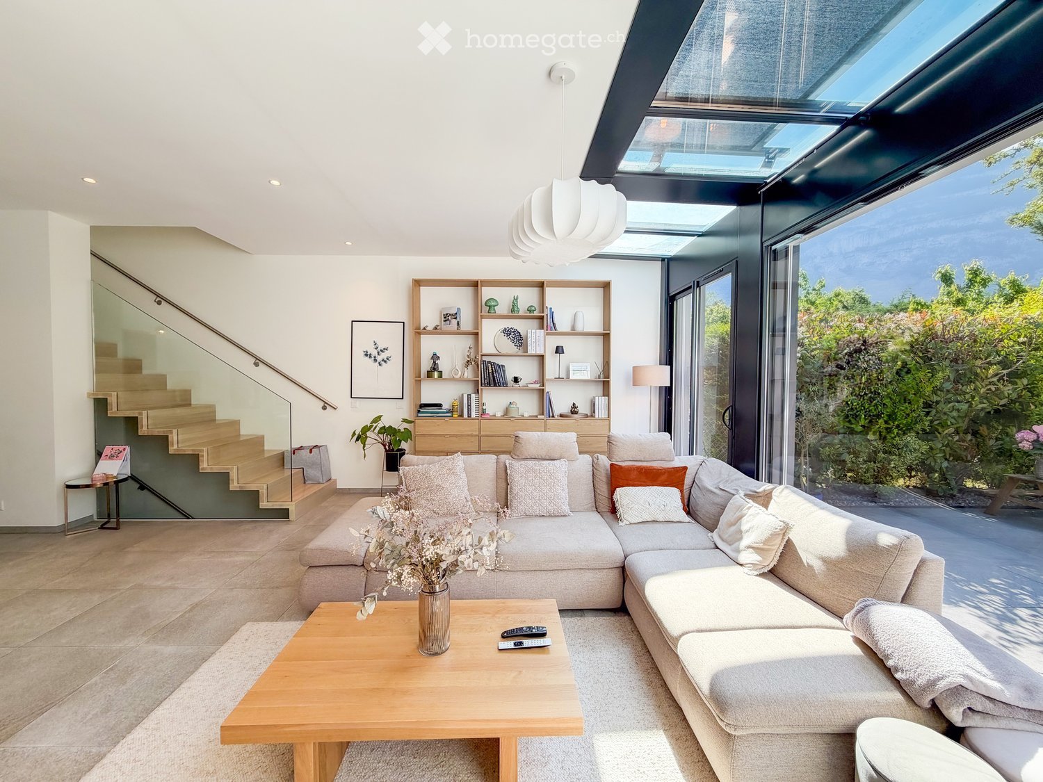 Modern living room with large windows and skylight, featuring a grey sectional sofa, wooden coffee table, bookshelf, and decorative items.
