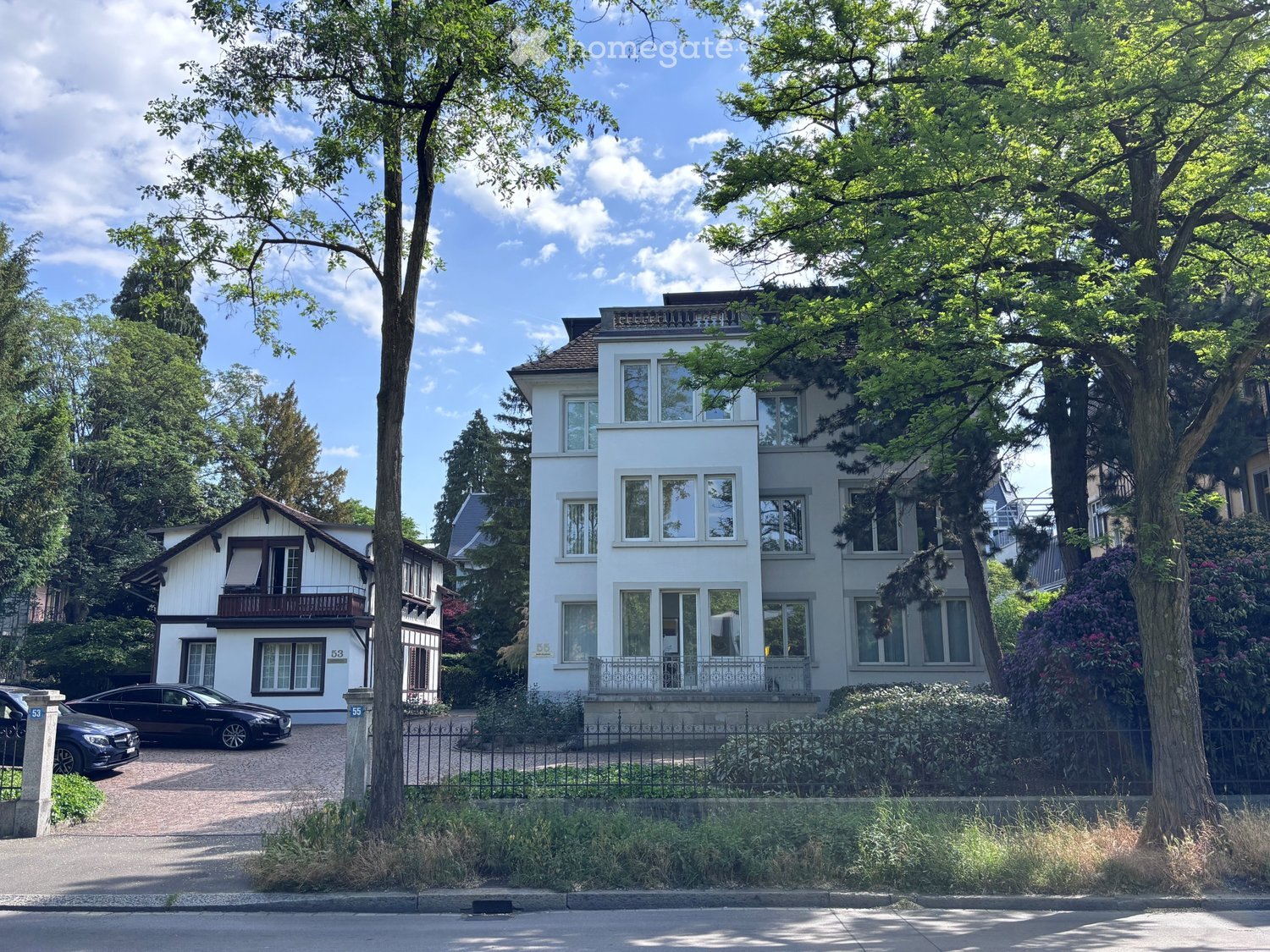 A two-story white house with large windows and a balcony, surrounded by trees and greenery, with a driveway and parked cars.