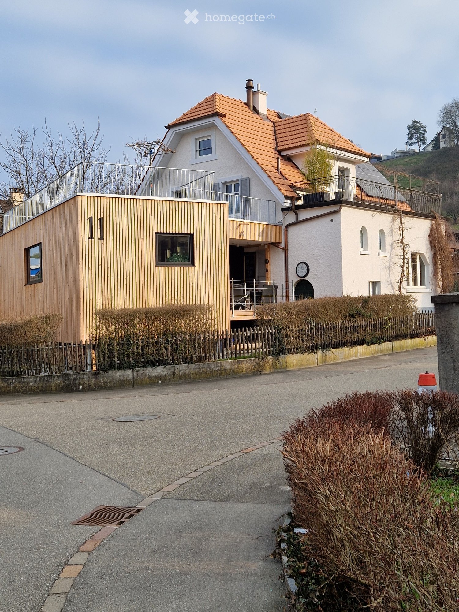 Two-story house with red-tiled roof, white walls, and a balcony. Adjacent wooden structure with large windows and a small patio area.