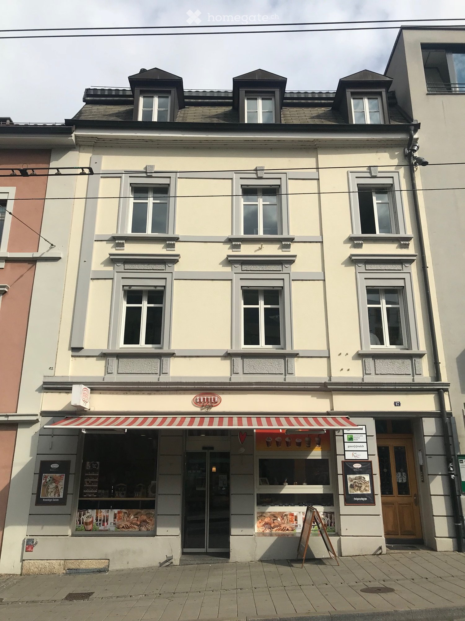 Three-story building with light yellow facade, grey trim, multiple windows, and a storefront with a red-and-white striped awning labeled "SUTTER".