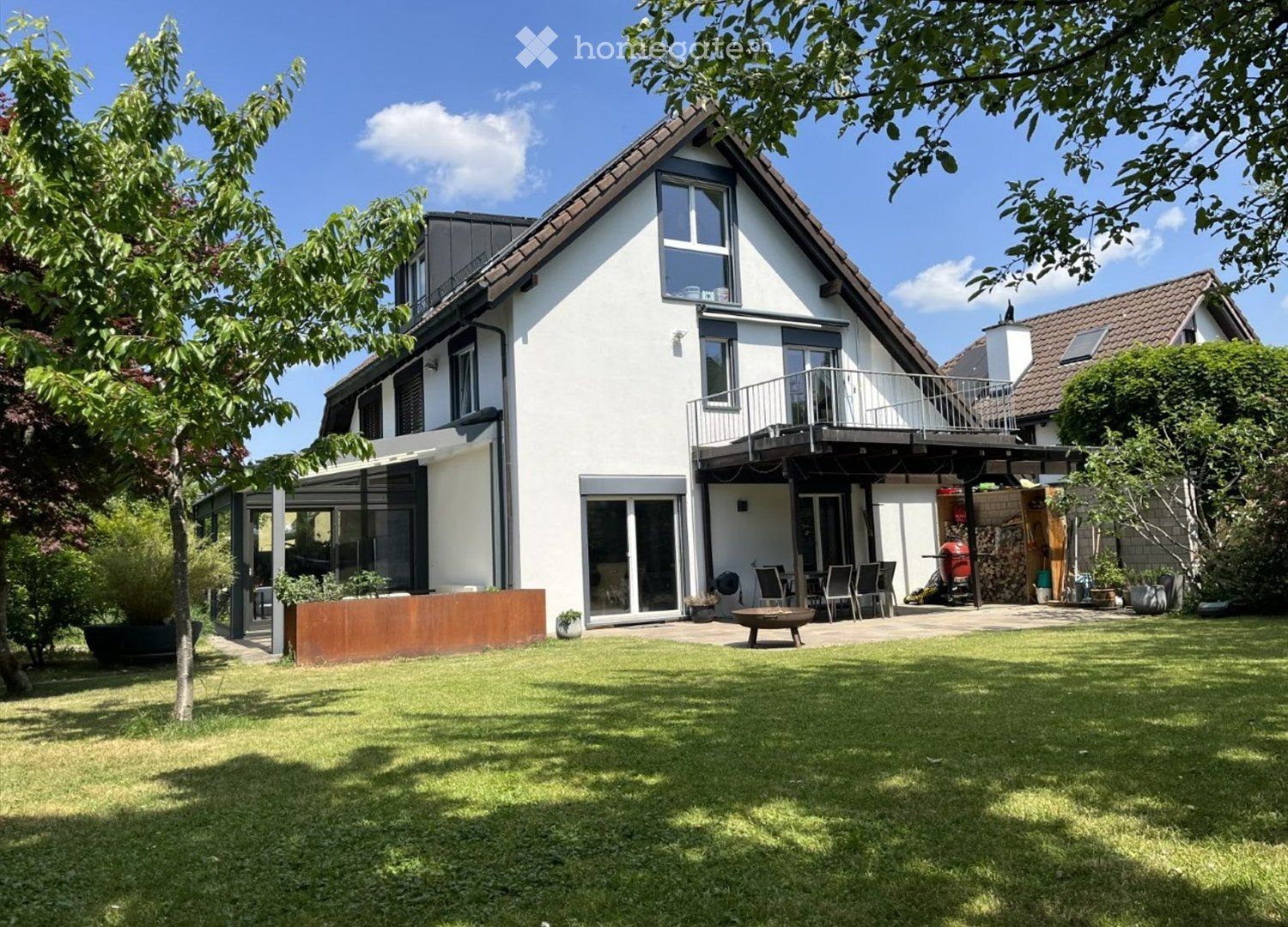 Two-story modern house with white exterior, large windows, and a balcony. The surrounding garden features a well-maintained lawn, patio seating area, and outdoor furniture.