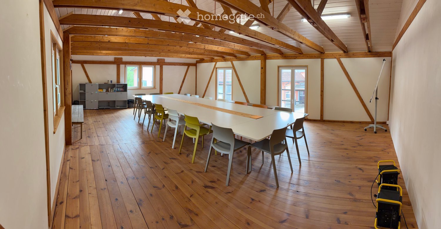 Bright, spacious room with wooden beams and polished wooden flooring, featuring a long white table surrounded by colorful chairs, a cabinet, and a stool.