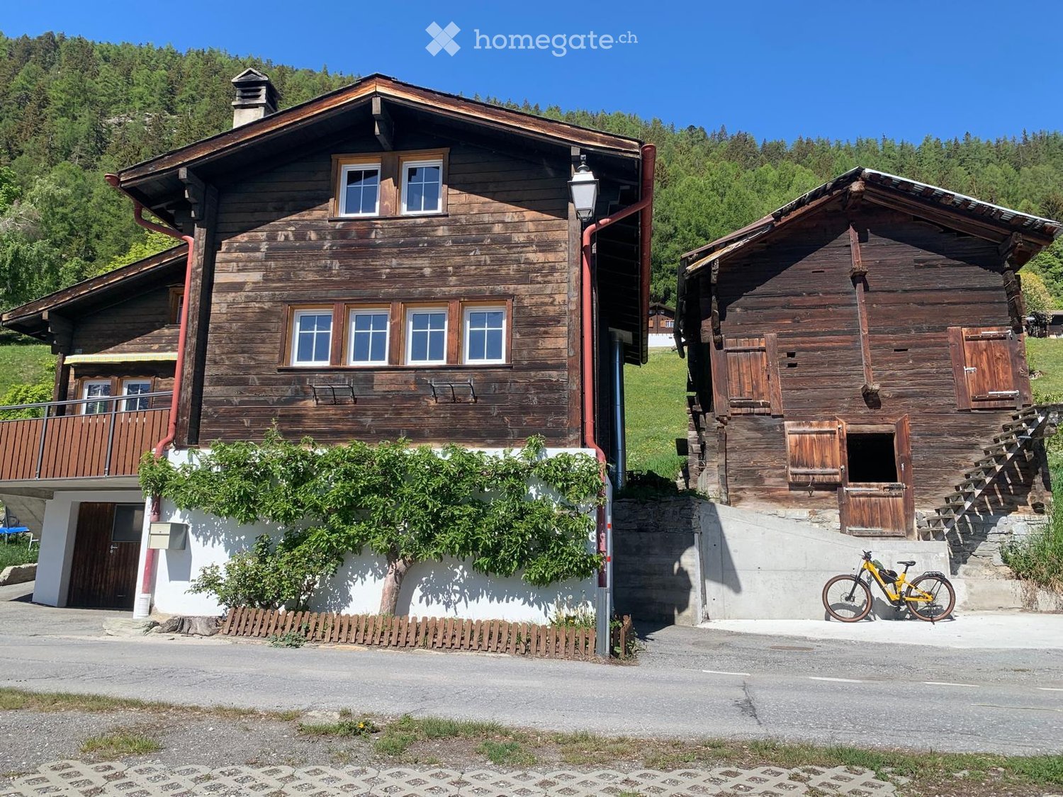 Two-story wooden house with multiple windows, a balcony, and a small shed. The house is surrounded by greenery and has a bicycle parked nearby.