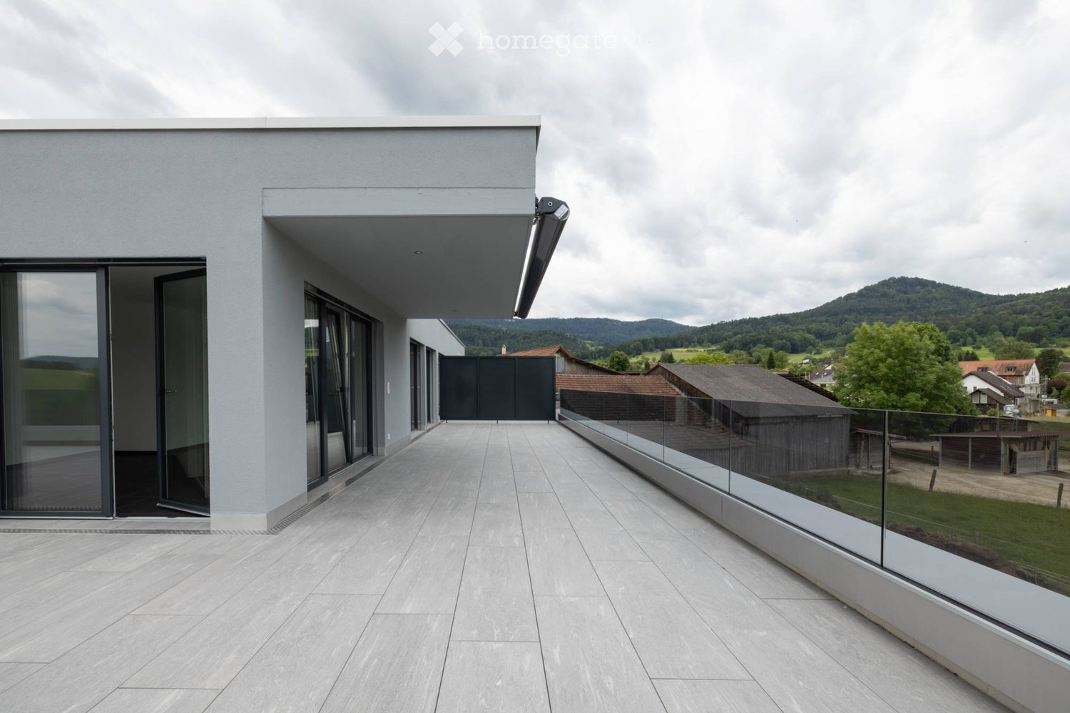 Modern balcony with large glass sliding doors, sleek grey tiles, and a glass railing overlooking a scenic countryside view.