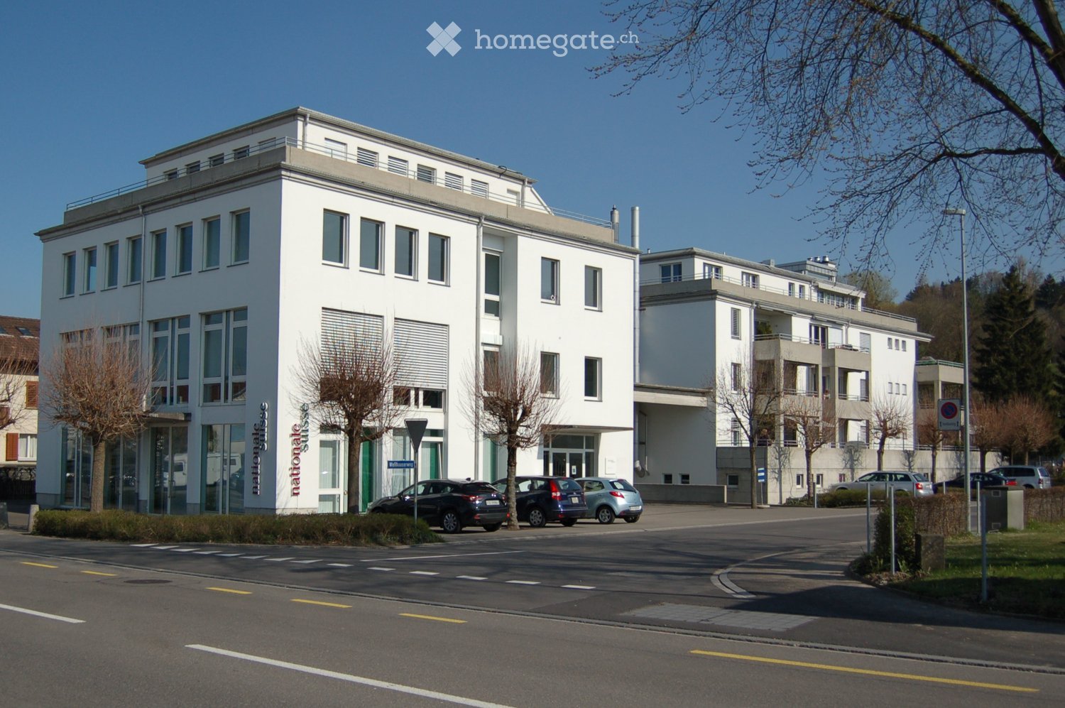 Modern building with multiple floors, white exterior, large windows, car parked in front, trees and greenery in front