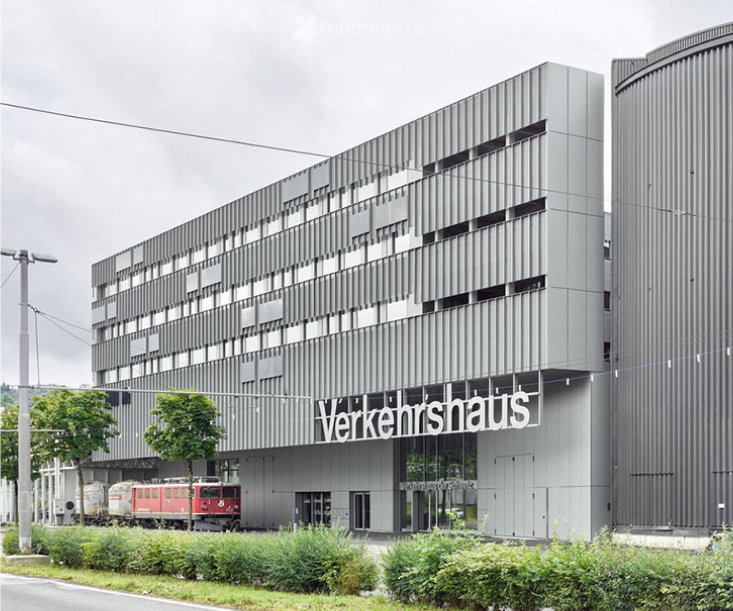 Modern, multi-story building with a sleek, metallic facade and large windows. The name "Verkehrshaus" is displayed prominently on the front. There is a train visible in the background, indicating proximity to a railway station.