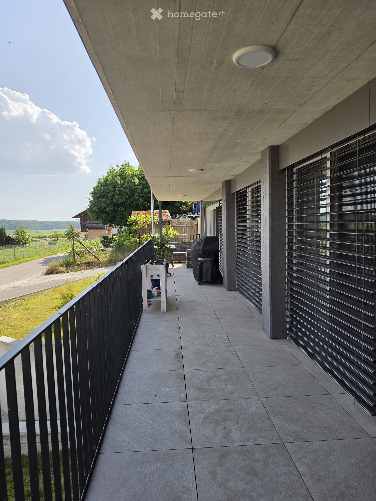 Modern balcony with concrete floor, black metal railing, and built-in blinds. Features a small table and a covered grill in the background.