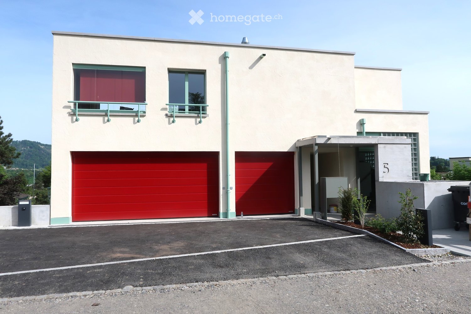 Modern two-story house with beige facade, red garage doors, and green-framed windows. Features a small balcony and a paved driveway.