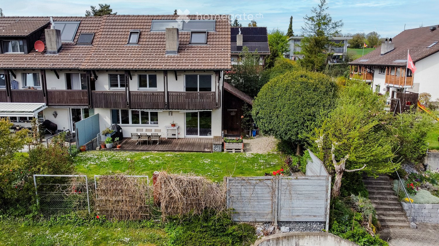 House. A multi-story house with a tiled roof, large windows, and balconies. The property includes a well-maintained garden with a patio area, outdoor furniture, and various plants.