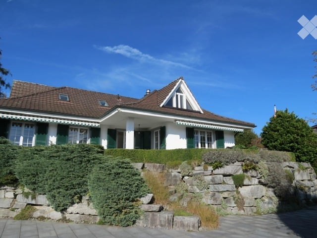 Two-story house with a brown tiled roof, white walls, green shutters, and a stone retaining wall in front.