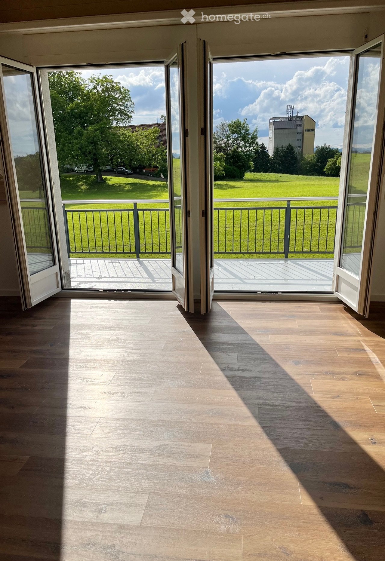 Empty room with wooden flooring and large open sliding doors leading to a balcony with a view of a green field and trees.