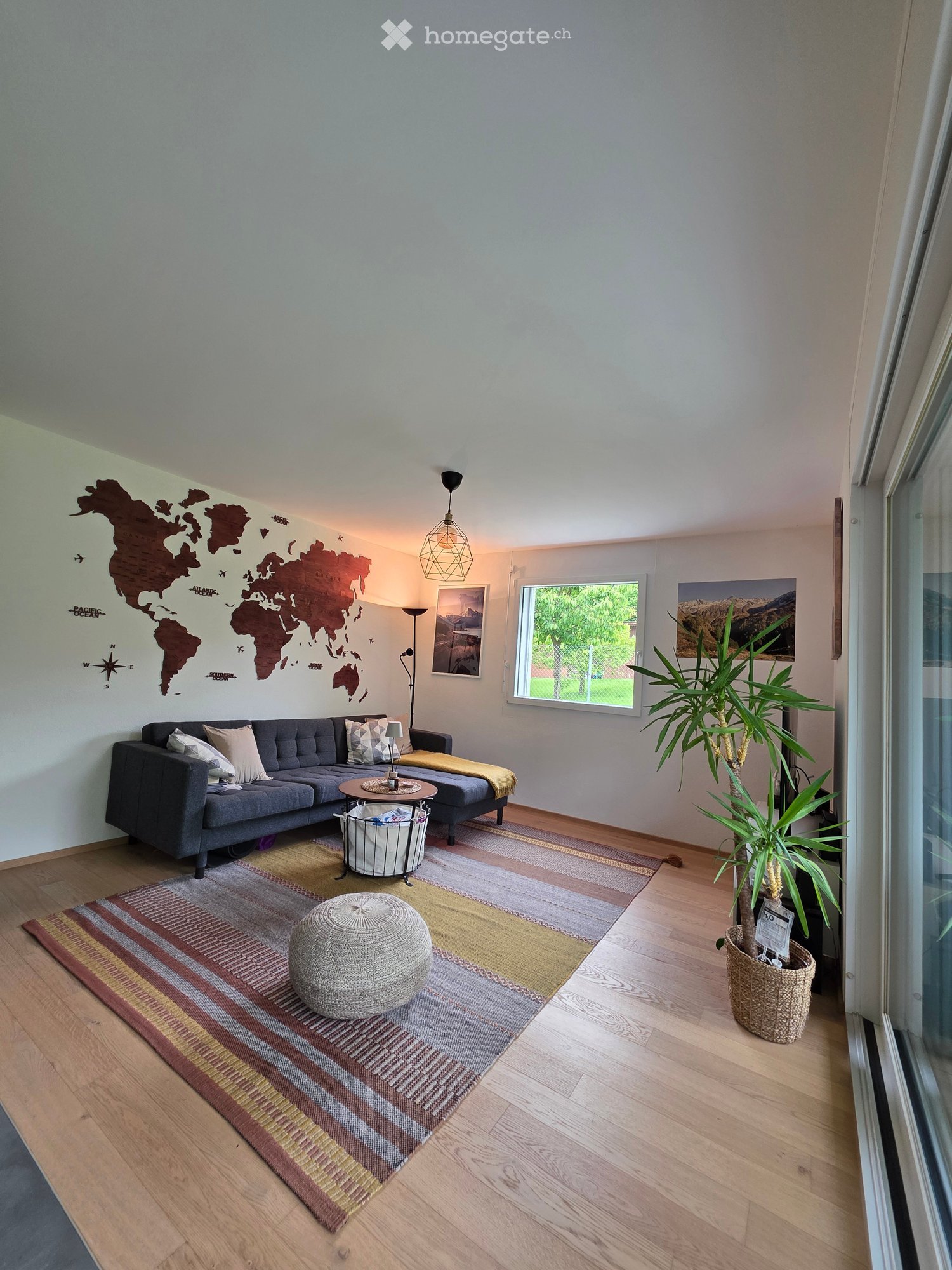 Modern living room with grey sectional sofa, coffee table, ottoman, floor lamp, potted plant, and world map wall art.