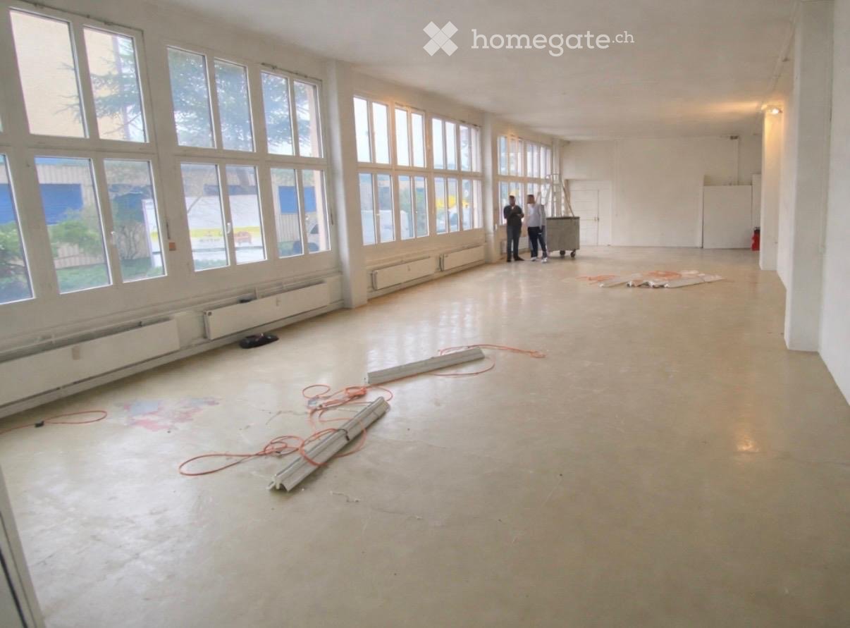 Empty room with large windows, concrete floor, and minimal furnishings. Two individuals are present, along with a dolly and some construction materials.