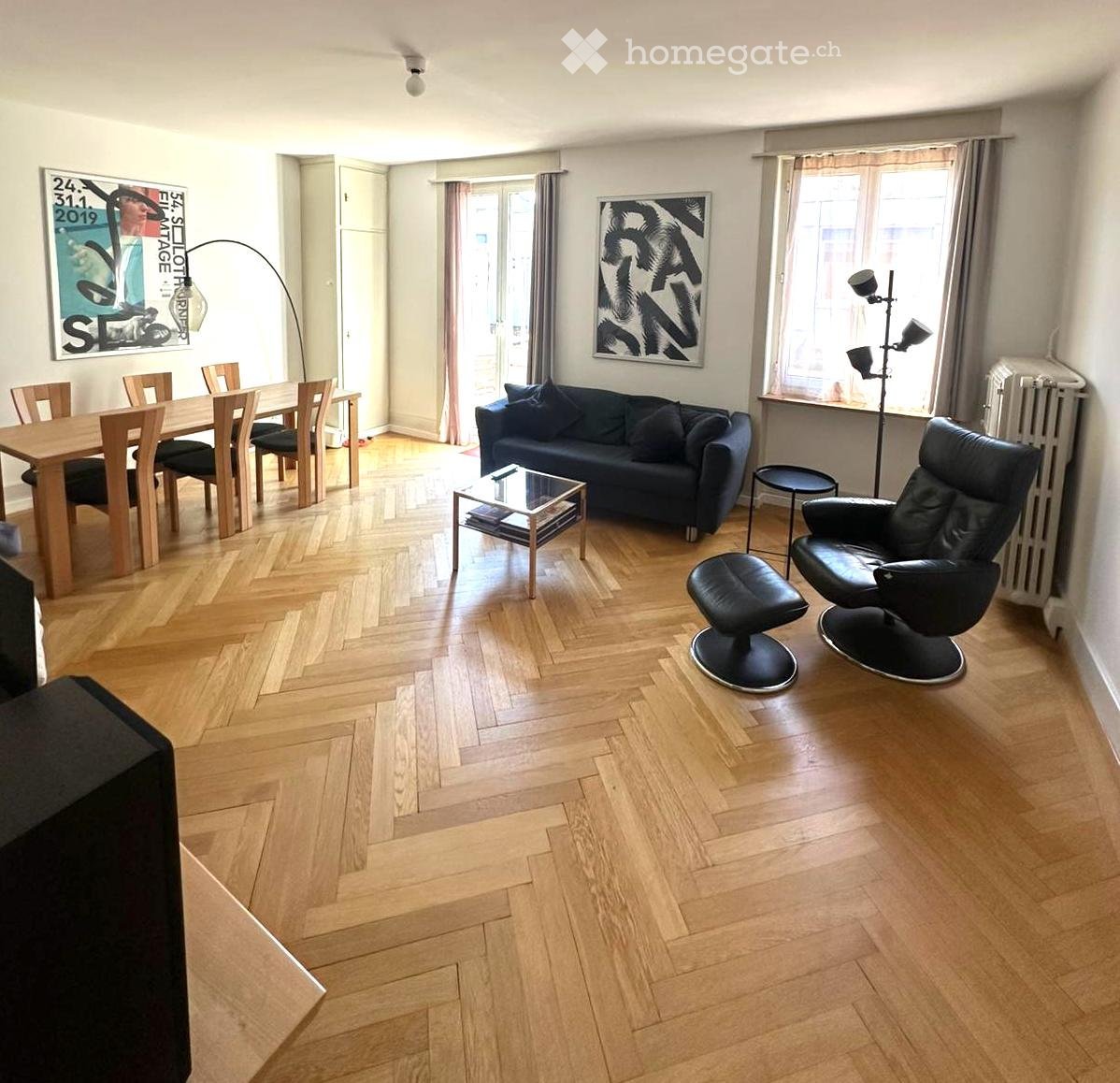 A modern, open-plan living and dining area with wooden herringbone flooring, a dark blue sofa, a glass coffee table, a wooden dining table with chairs, and a black leather armchair.