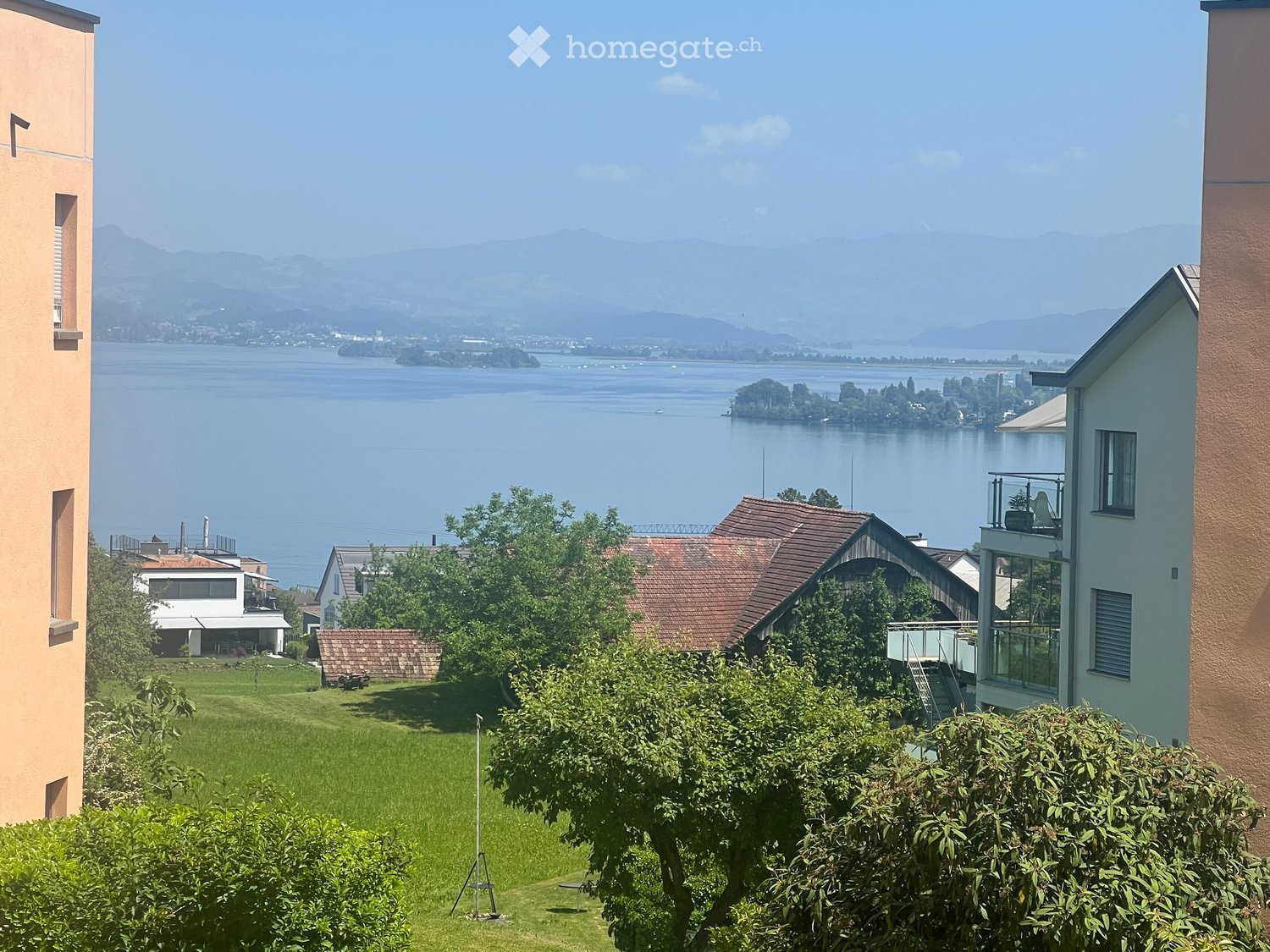 View of a lake and distant mountains from a garden area with green grass and trees, surrounded by residential buildings.