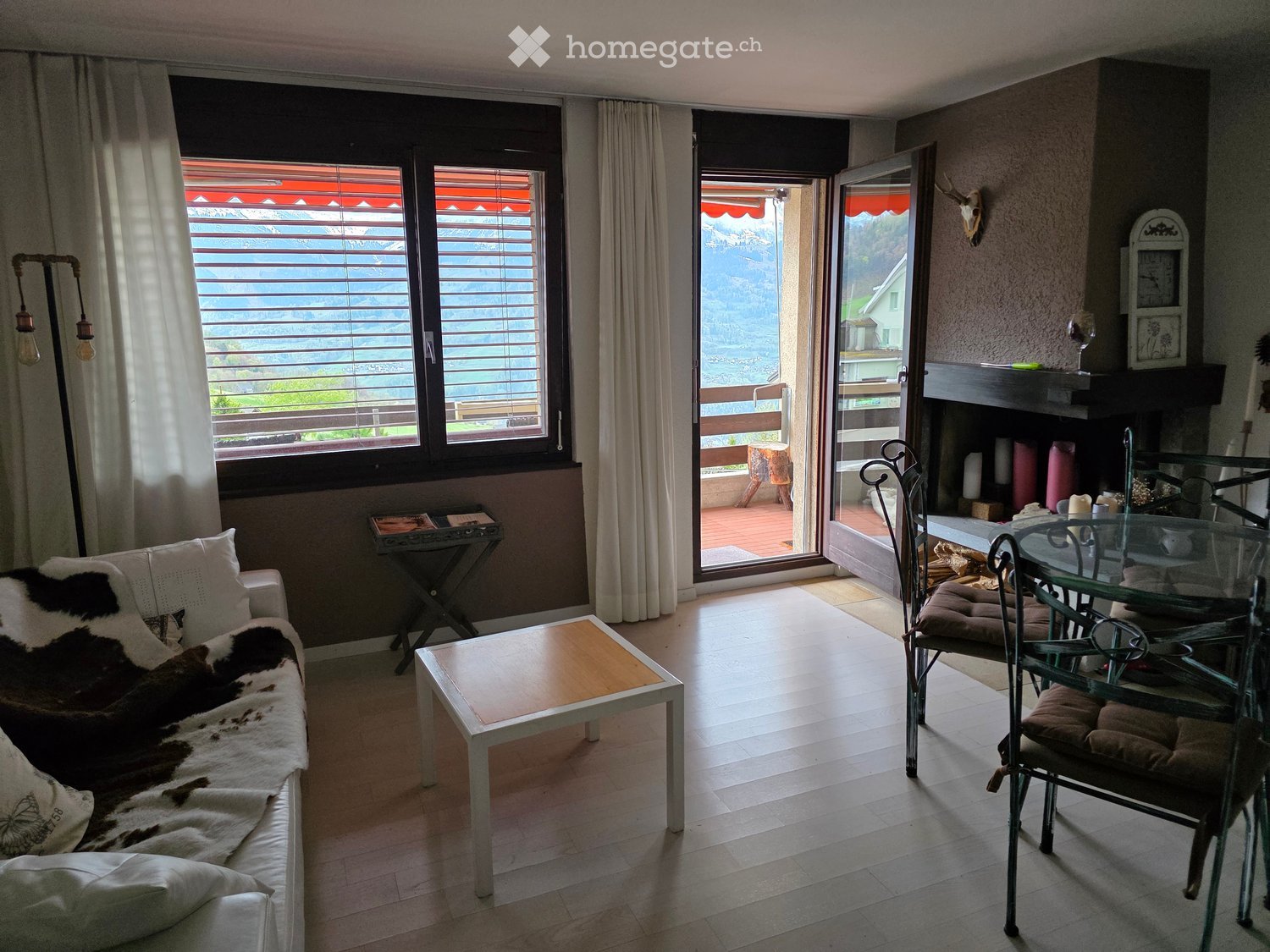 Bright living room with large windows and a balcony overlooking a scenic mountain view. Features a white sofa with a cowhide throw, a wooden coffee table, a glass dining table with chairs, and a fireplace with decorative items.