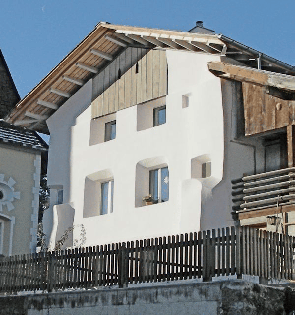 A modern white house with a wooden roof and multiple windows, featuring a small balcony with a flower pot and a wooden fence in front.