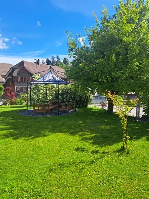Well-maintained garden with a gazebo, wooden benches, and lush greenery, featuring a house in the background.