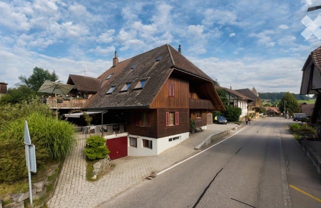Traditional Swiss wooden house with a steeply pitched roof, red shutters, and a red garage door, situated in a picturesque village setting with other houses and greenery.