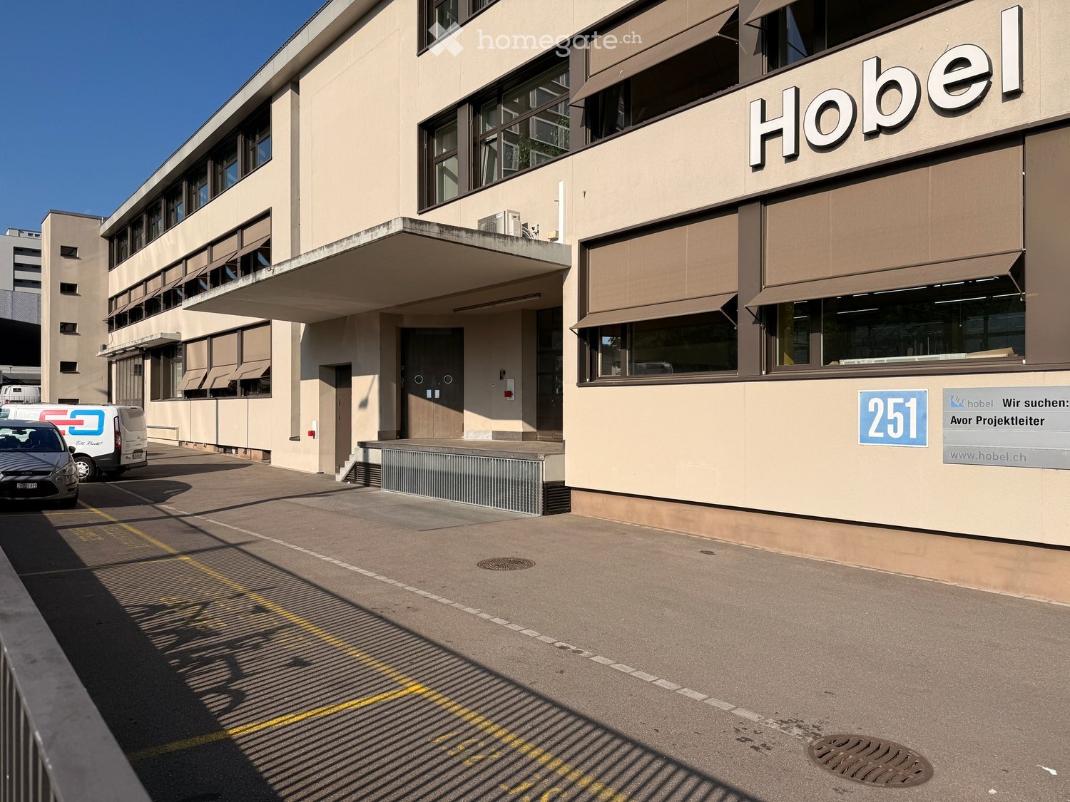 Modern multi-story building with beige facade, large windows, and a sign reading "Hobel" on the upper right. The entrance features a covered area with a door and a small staircase leading up. A parking area is visible in front with a few cars and a van.