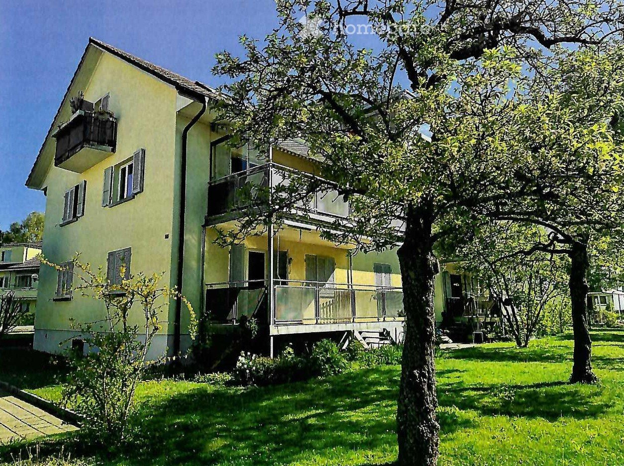 Two-story yellow house with balconies, surrounded by green lawn and trees.
