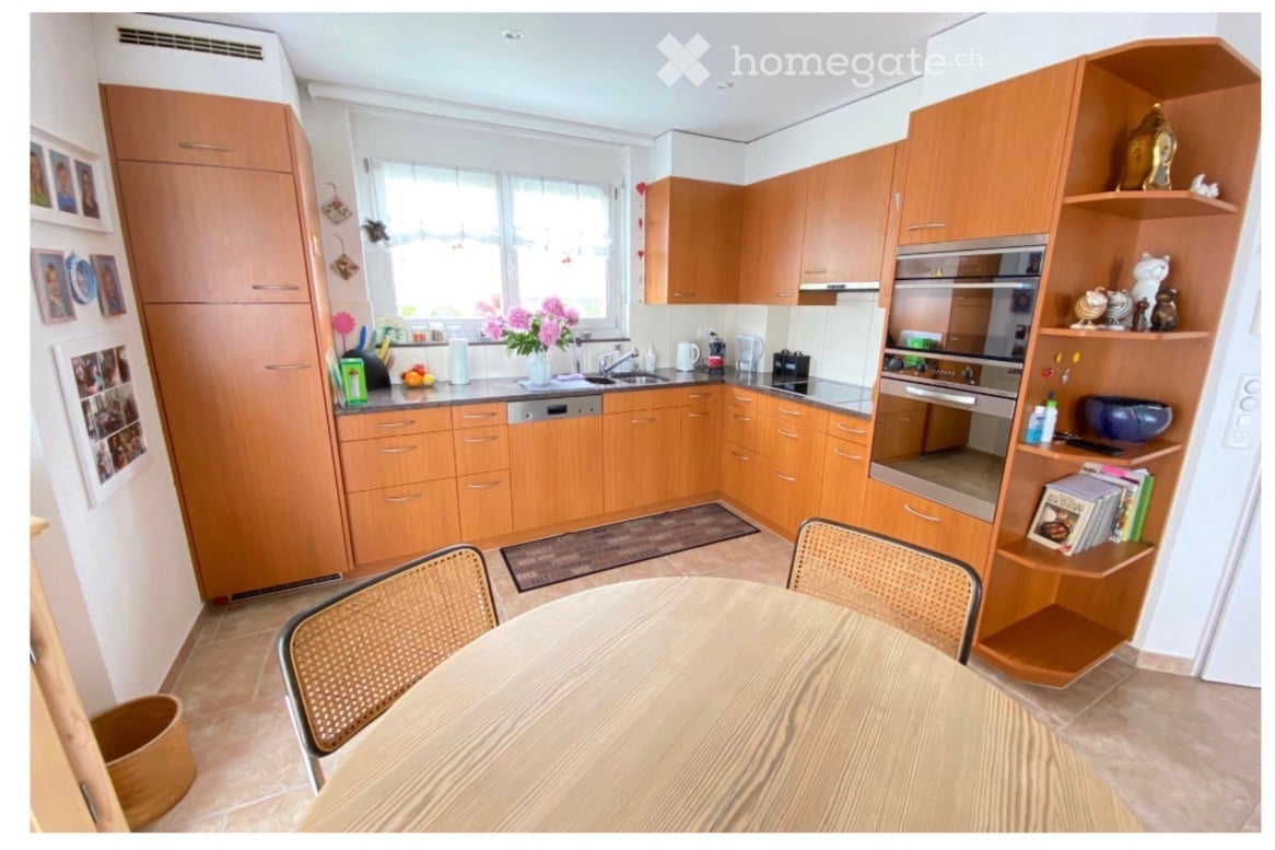 A modern kitchen with wooden cabinets, a round wooden dining table with two chairs, a built-in oven, a dishwasher, and a shelf with decorative items and books.