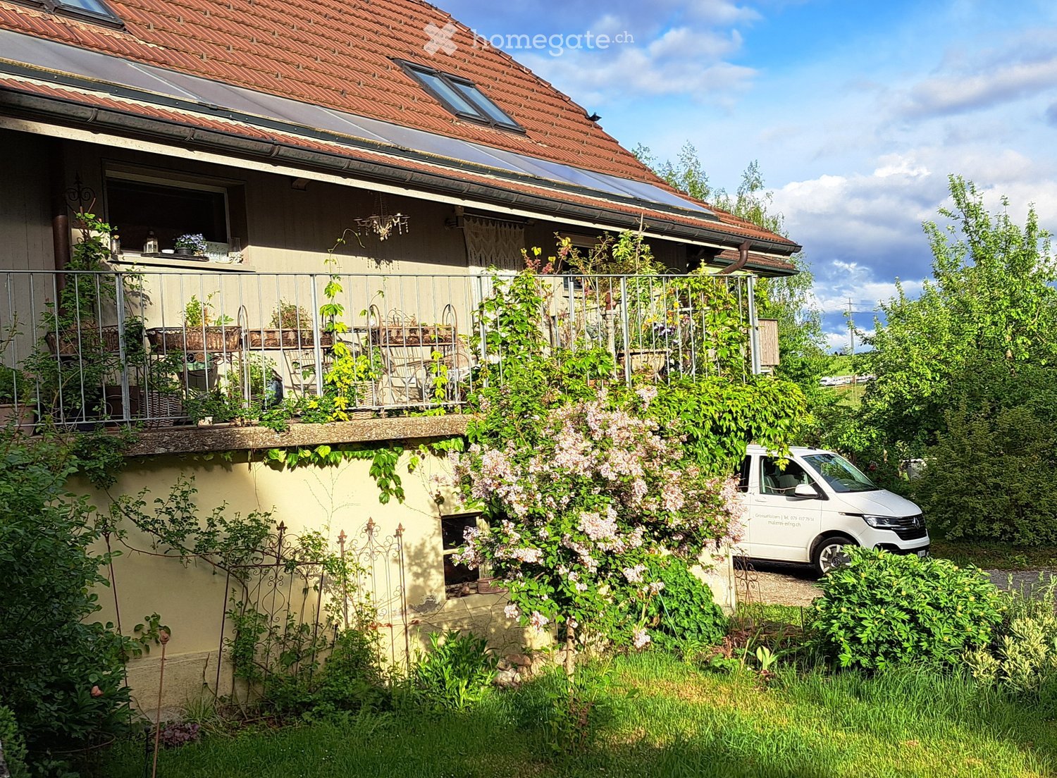 Two-story house with red-tiled roof, solar panels, balcony, and lush greenery surrounding the property.