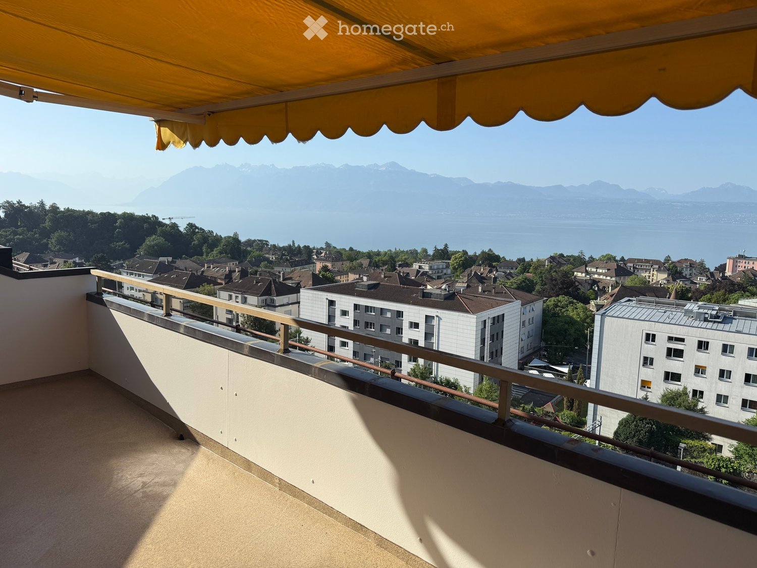 Balcony with metal railing, yellow awning, scenic view of city, lake, and mountains.