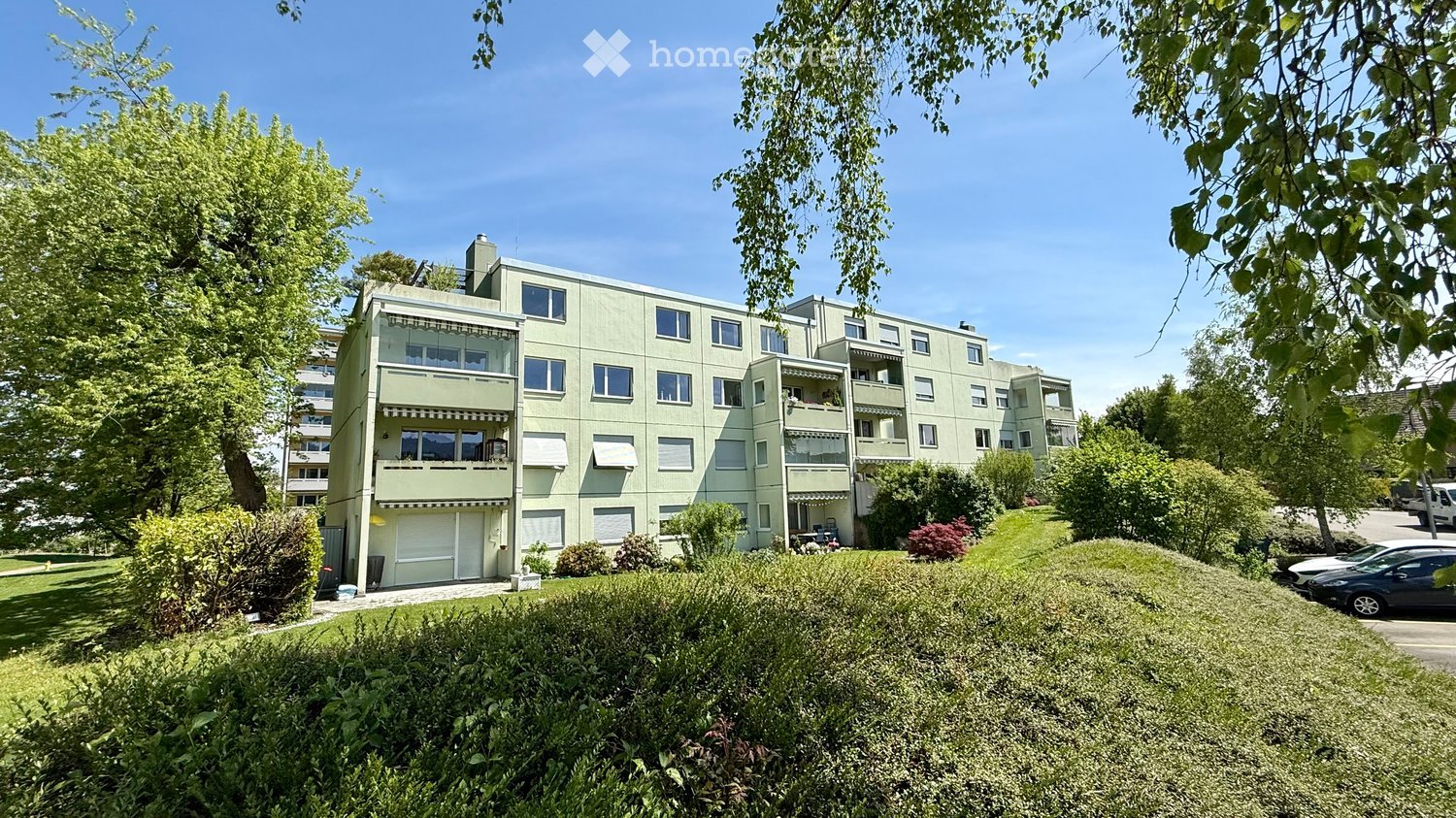 A multi-story residential building with light green facade, multiple windows, balconies, and surrounding greenery.