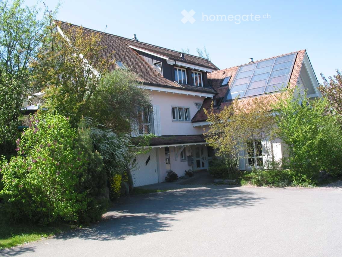 Two-story house with a red-tiled roof, light pink walls, multiple windows, and a driveway surrounded by greenery.