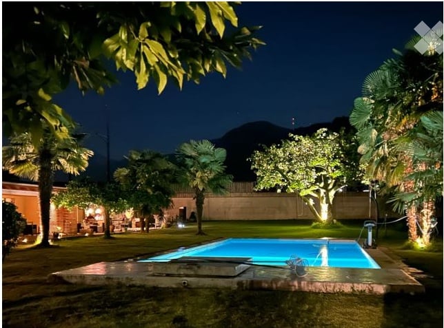Nighttime view of a well-maintained garden with a swimming pool, surrounded by palm trees and other greenery, illuminated by ambient lighting.
