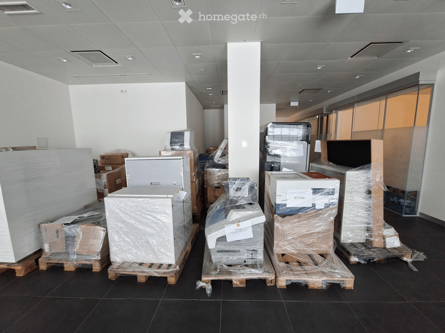 Empty room with black tiled floor, white walls, and several large appliances and equipment wrapped in plastic and placed on pallets.