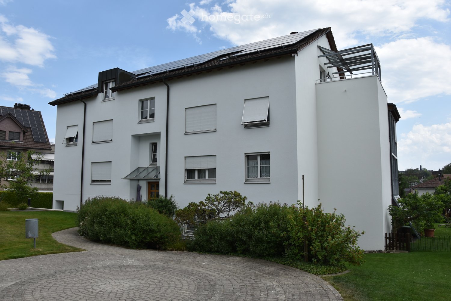 Modern two-story white house with solar panels on the roof, multiple windows with shutters, a small balcony, and a landscaped garden with a paved driveway.