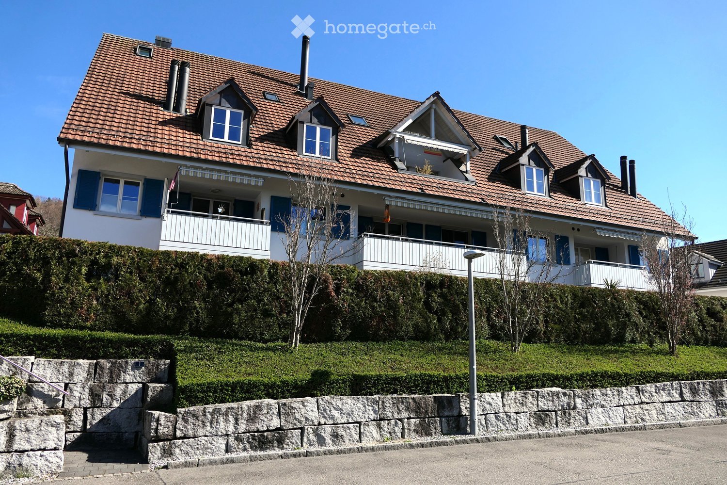 3 story house, brown roof, blue shutters, multiple balconies, stone wall, hedges, street lamp